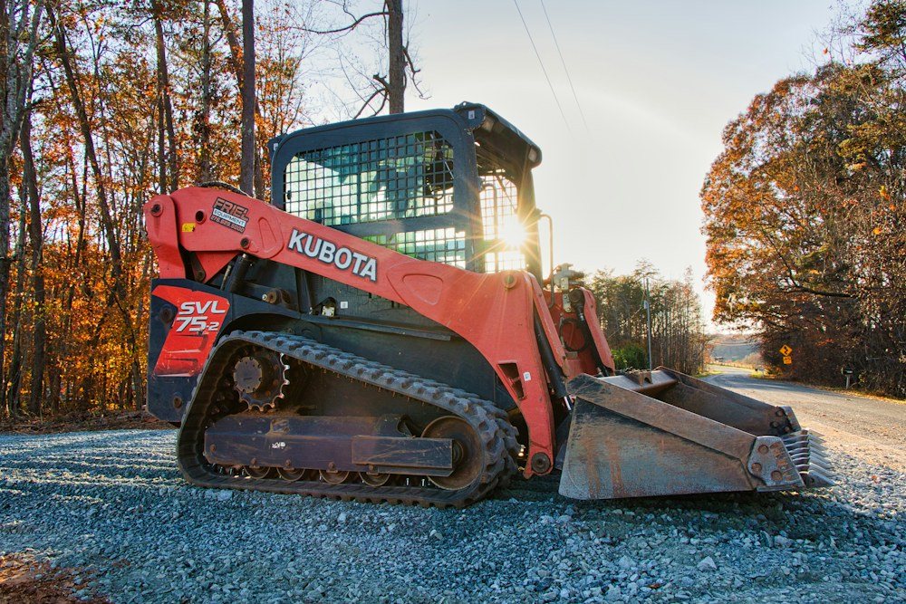 A compact track loader parked at the edge of a field beside a hardwood tree line